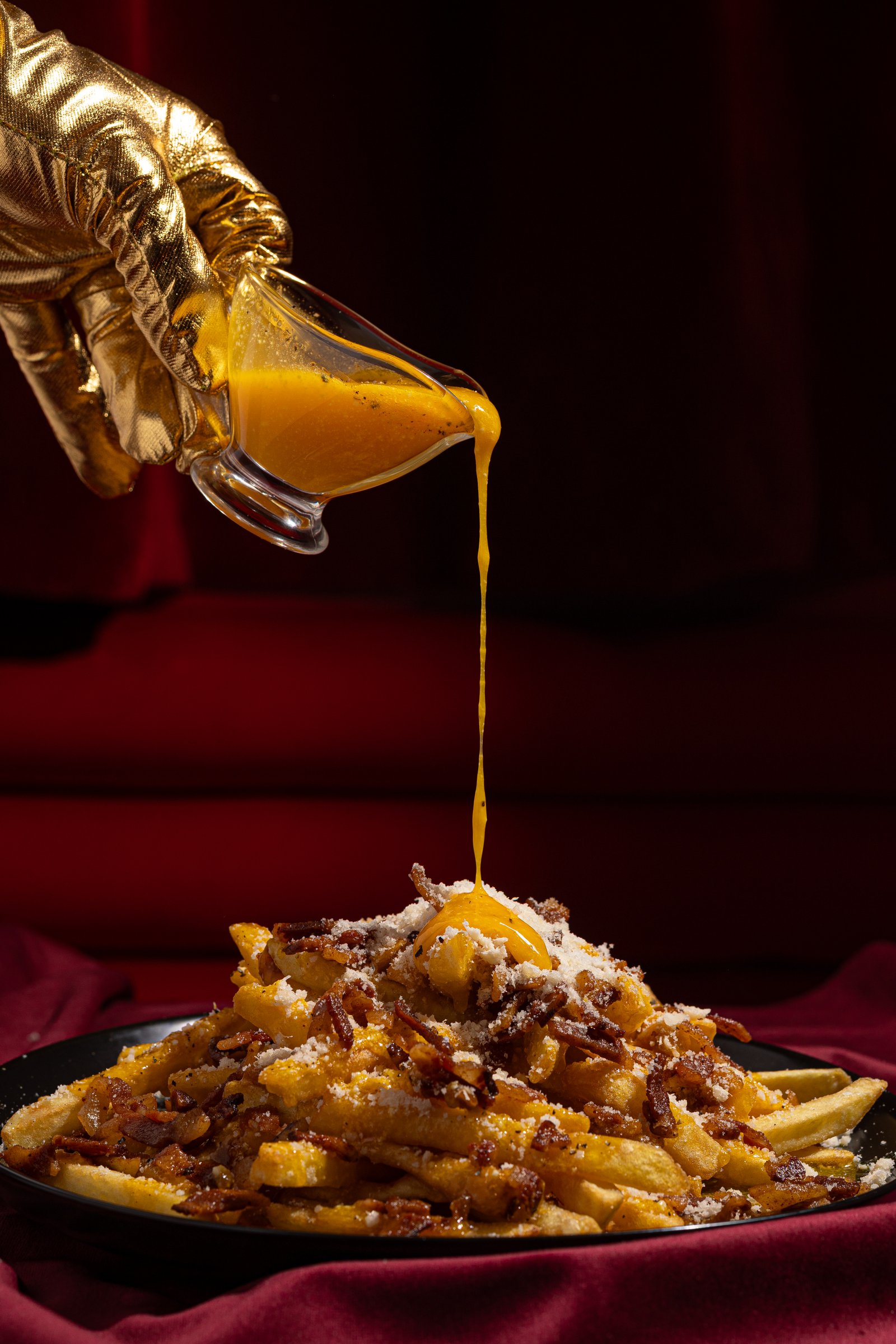 Mano enguantada en oro vertiendo salsa cheddar dorada sobre patatas con bacon — Burgaccia