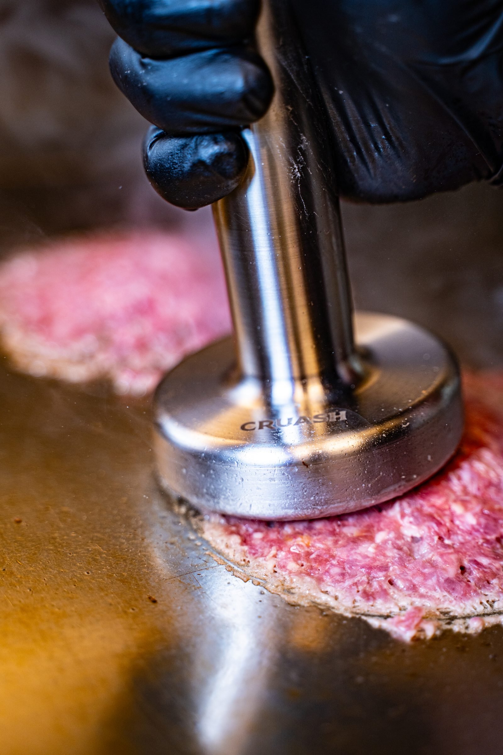 Smasher CRUASH de acero con marca grabada presionando una hamburguesa de carne fresca sobre la plancha — fotografía de producto en uso