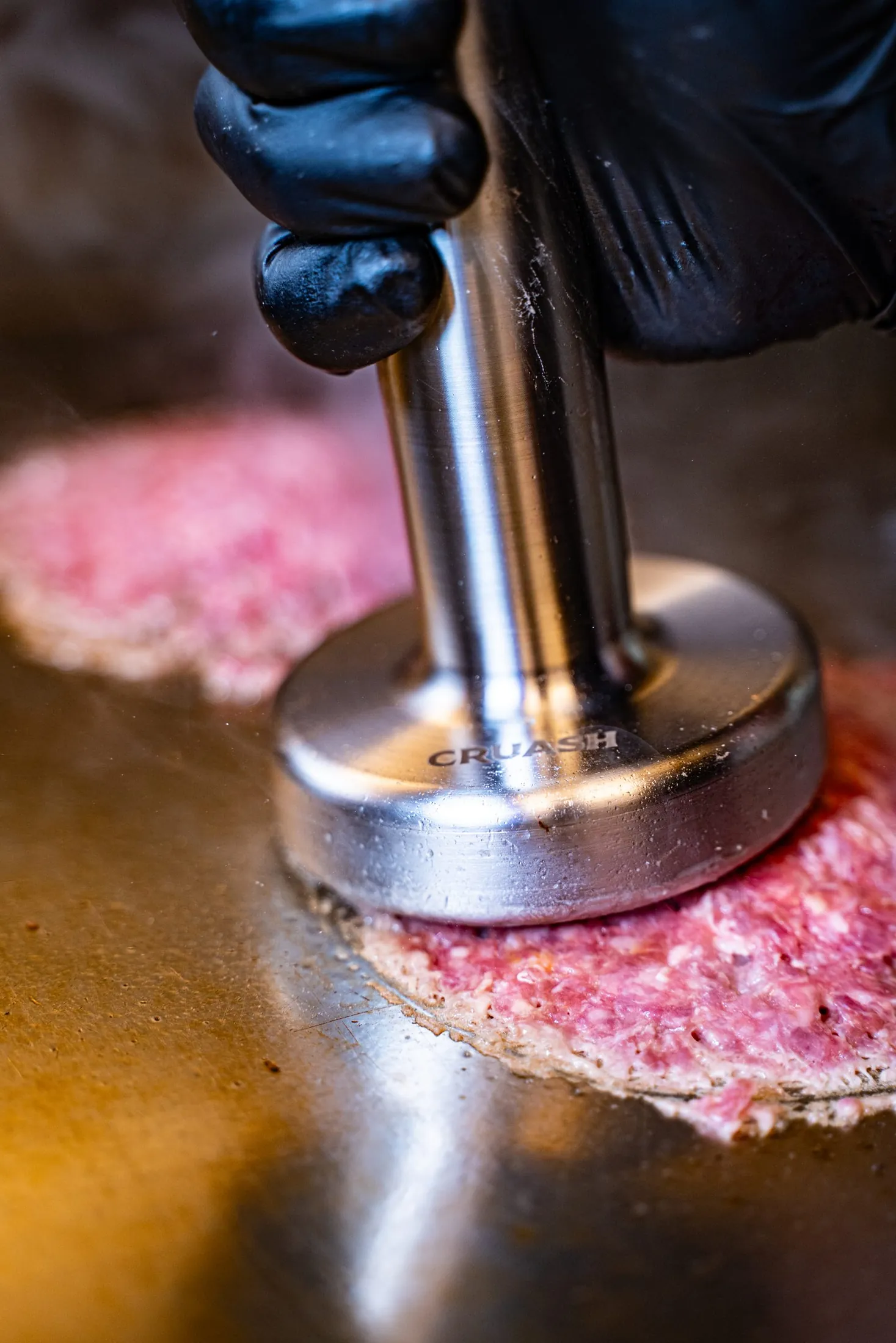 Smasher CRUASH de acero con marca grabada presionando una hamburguesa de carne fresca sobre la plancha — fotografía de producto en uso