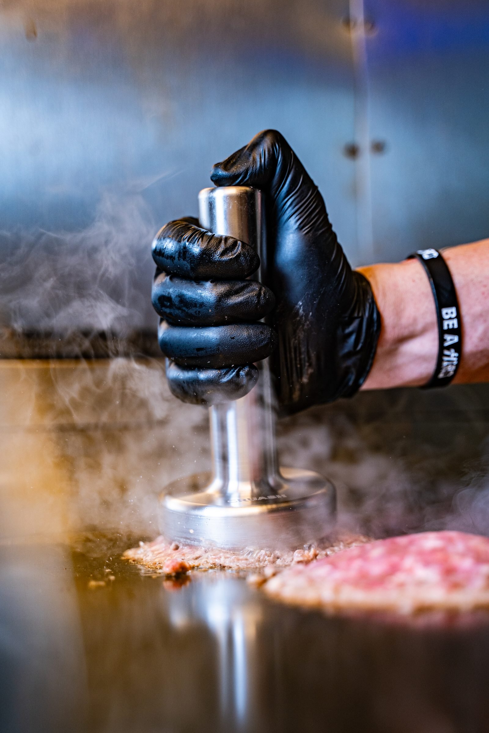 Mano con guante de nitrilo negro presionando el smasher CRUASH sobre una bola de carne mientras sale humo de la plancha — sesión editorial