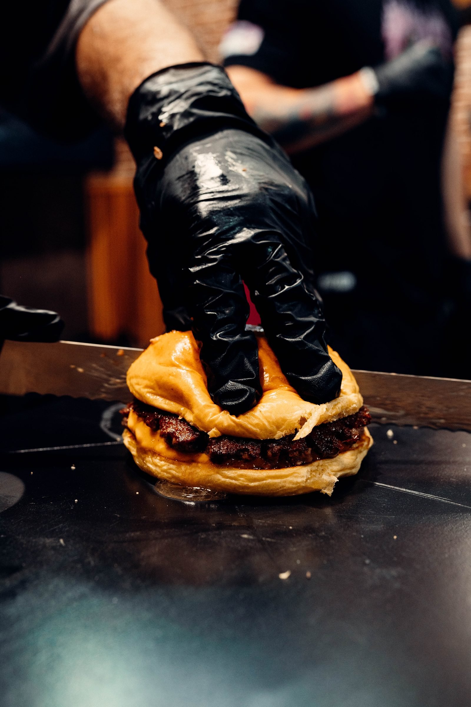 Mano con guante negro presionando un smashburger con queso fundido sobre la plancha — sesión CRUASH