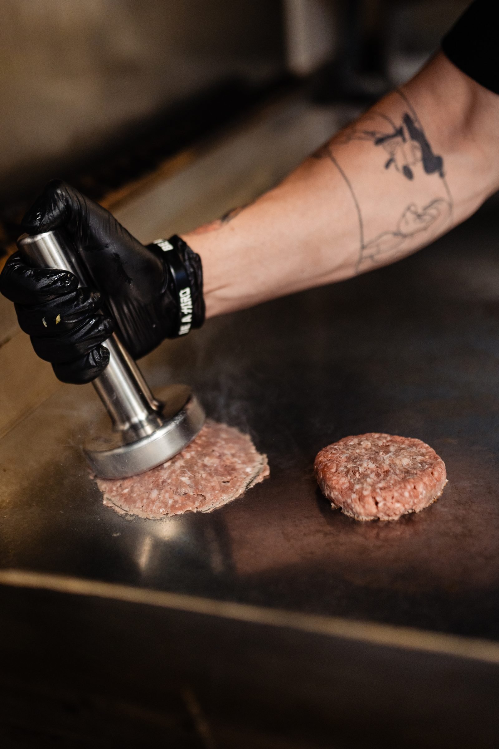 Plancha con dos smash burgers en distintos estadios de cocción mientras el smasher CRUASH presiona uno de ellos