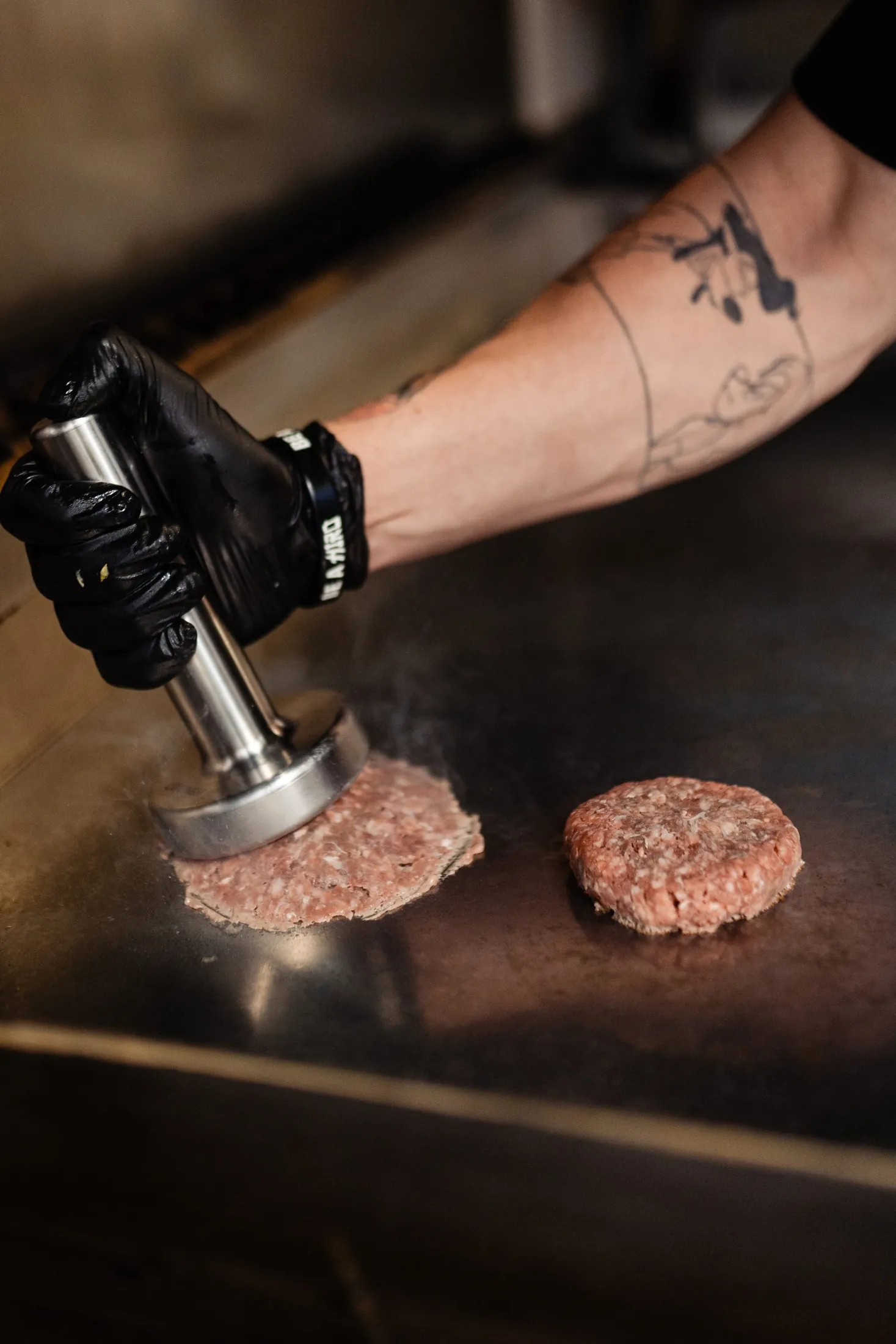 Plancha con dos smash burgers en distintos estadios de cocción mientras el smasher CRUASH presiona uno de ellos