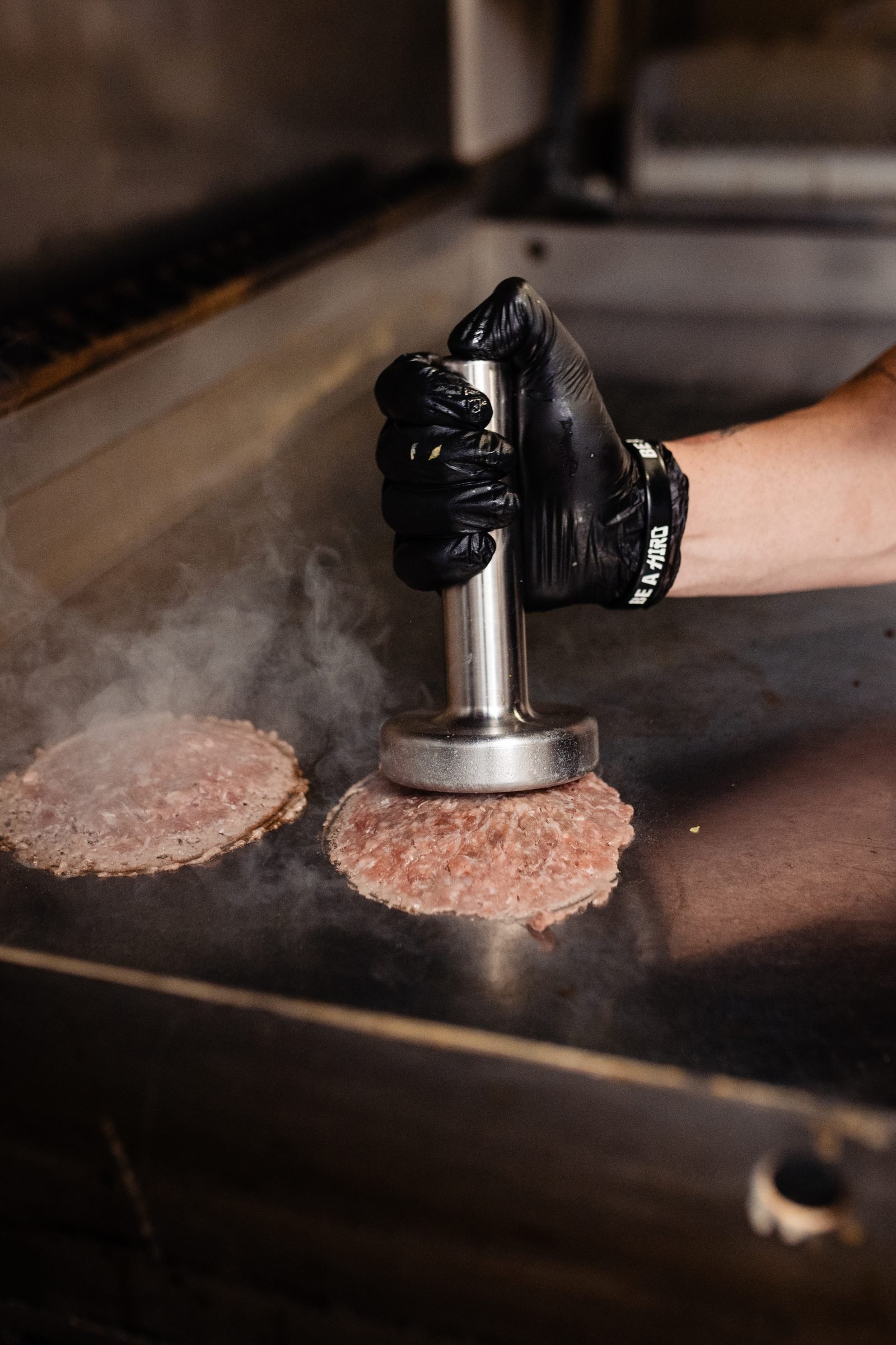 Vapor saliendo de la plancha mientras el smasher CRUASH aplasta una smash burger — fotografía cinemática