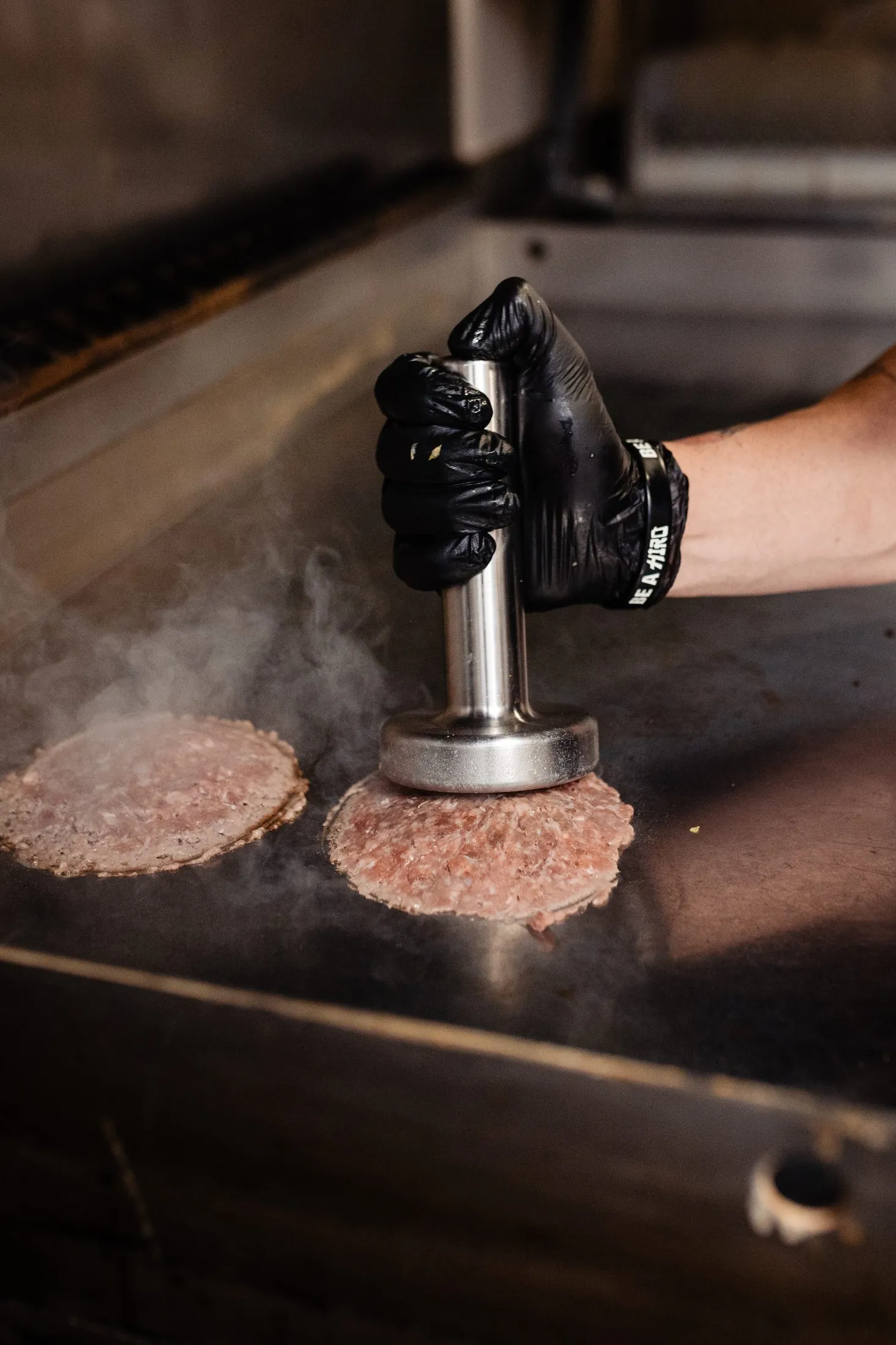 Vapor saliendo de la plancha mientras el smasher CRUASH aplasta una smash burger — fotografía cinemática