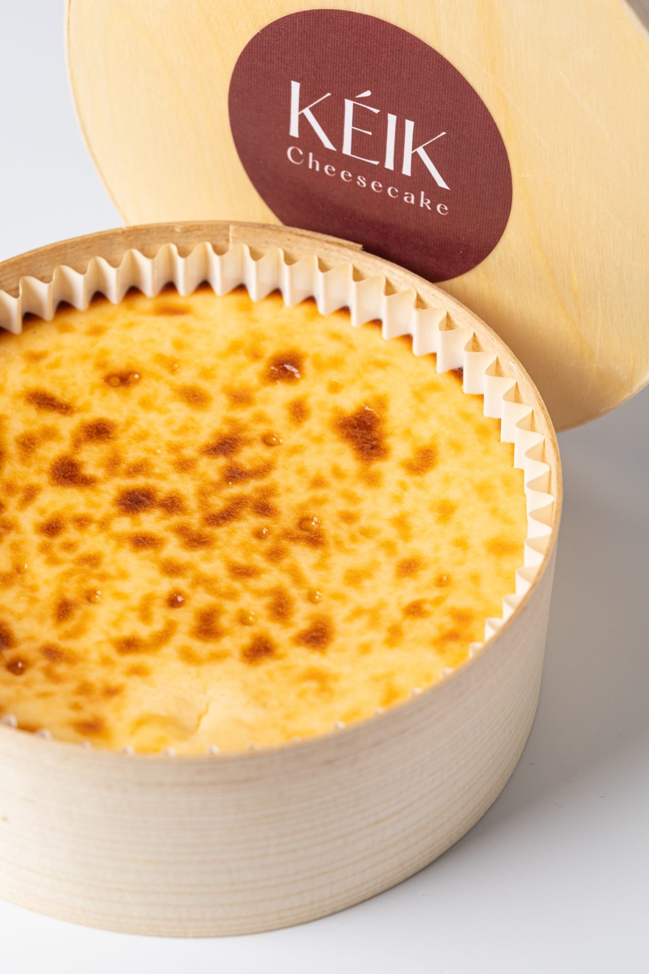 Tarta de queso vasca tostada en caja de madera circular con etiqueta KÉIK Cheesecake — fotografía de producto sobre fondo blanco