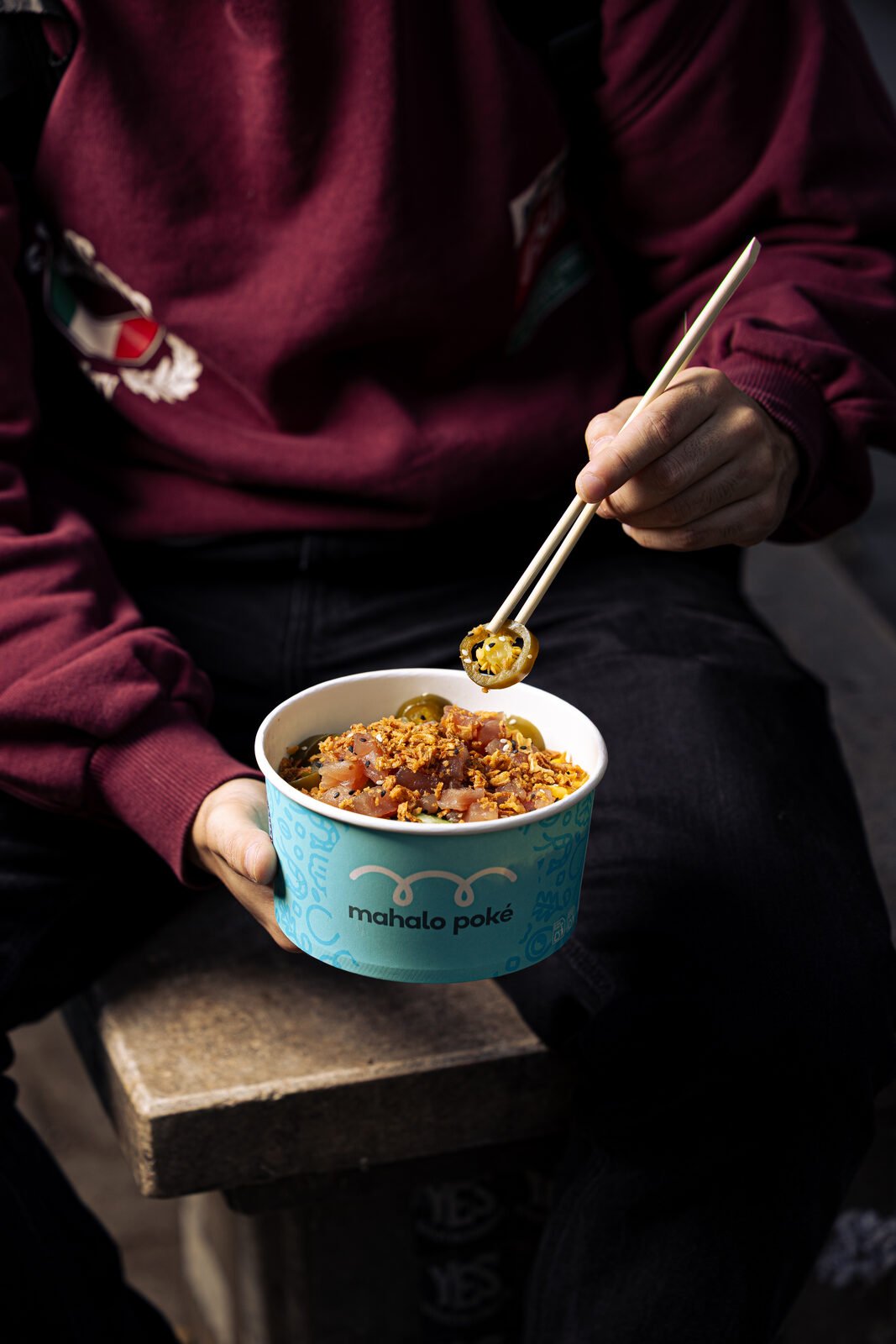 Persona con sudadera burdeos sosteniendo un bowl Mahalo Poké en la calle