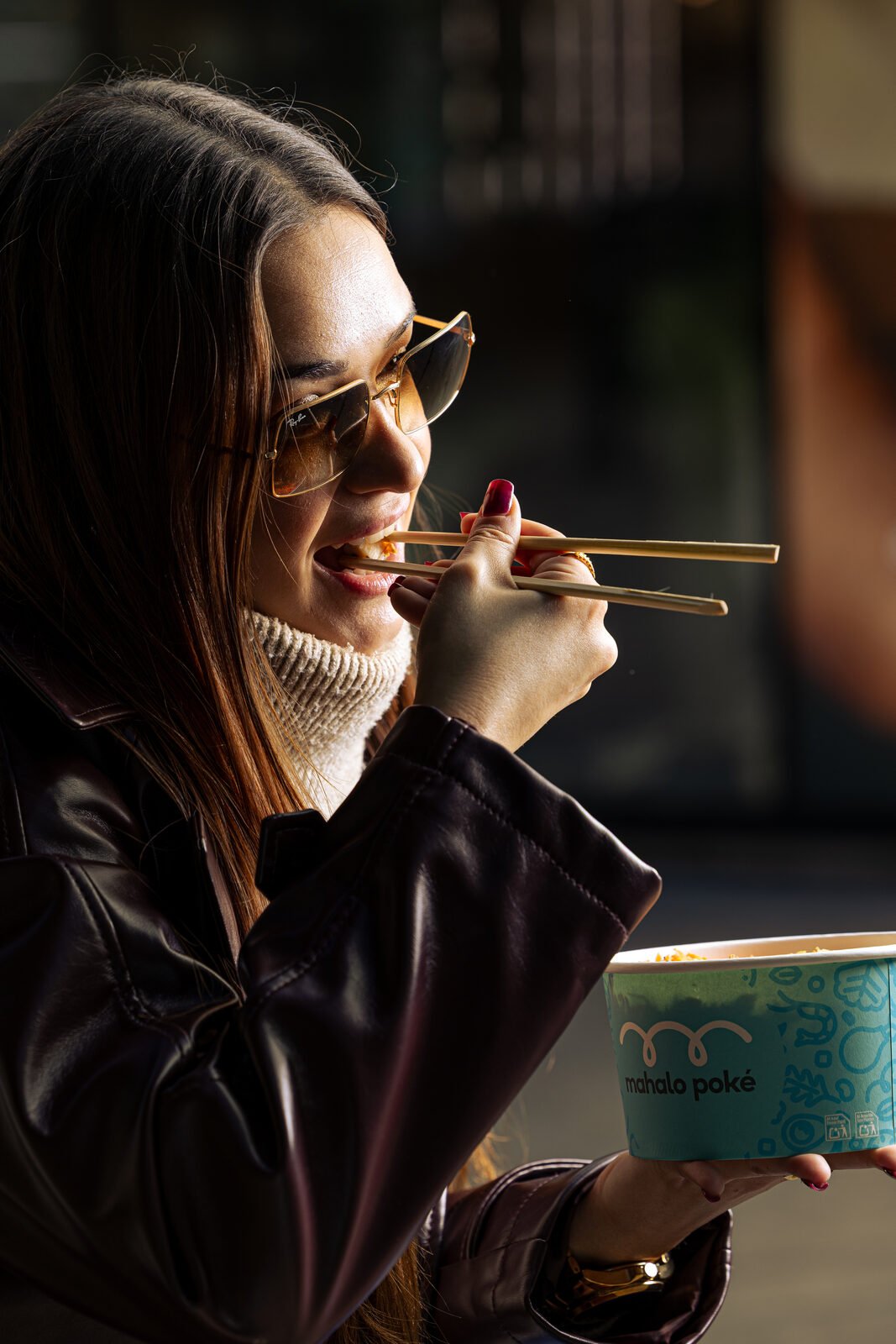 Mujer con chaqueta de cuero y gafas de sol comiendo poké de Mahalo a contraluz