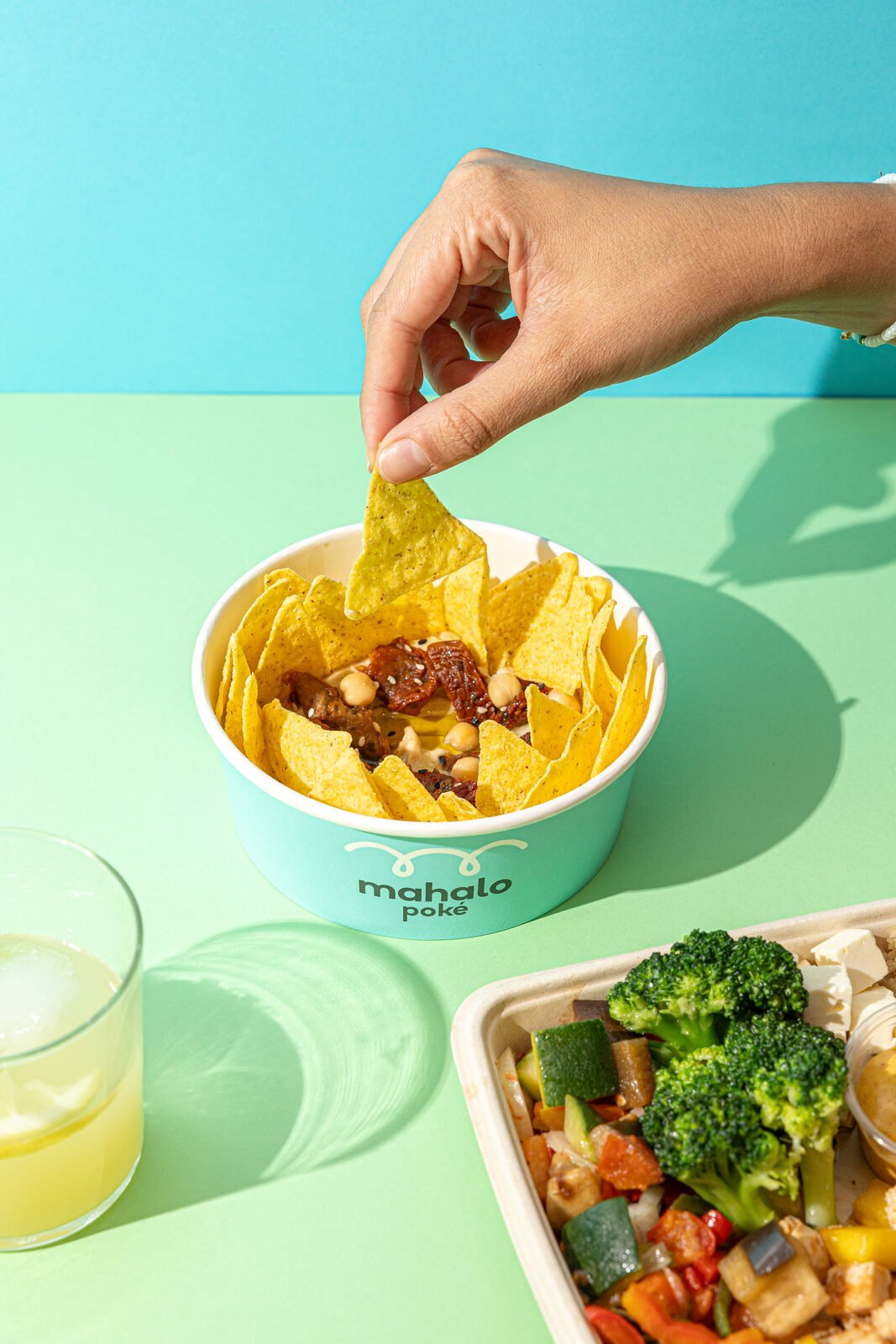 Mano sujetando un nacho sobre bowl de Mahalo Poké con fondo bicolor azul y verde menta