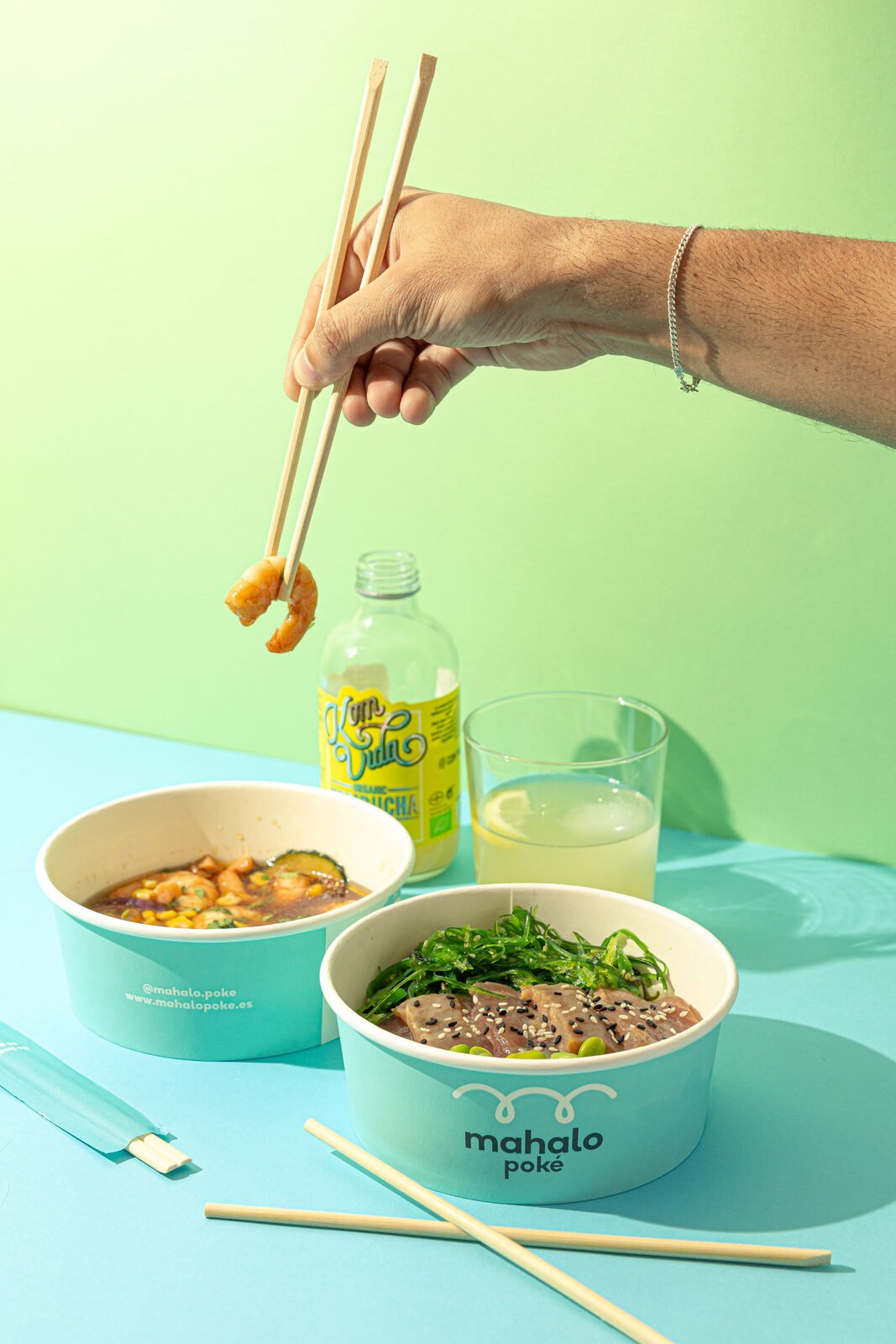 Mano con palillos levantando un langostino sobre bowls Mahalo y kombucha Komvida