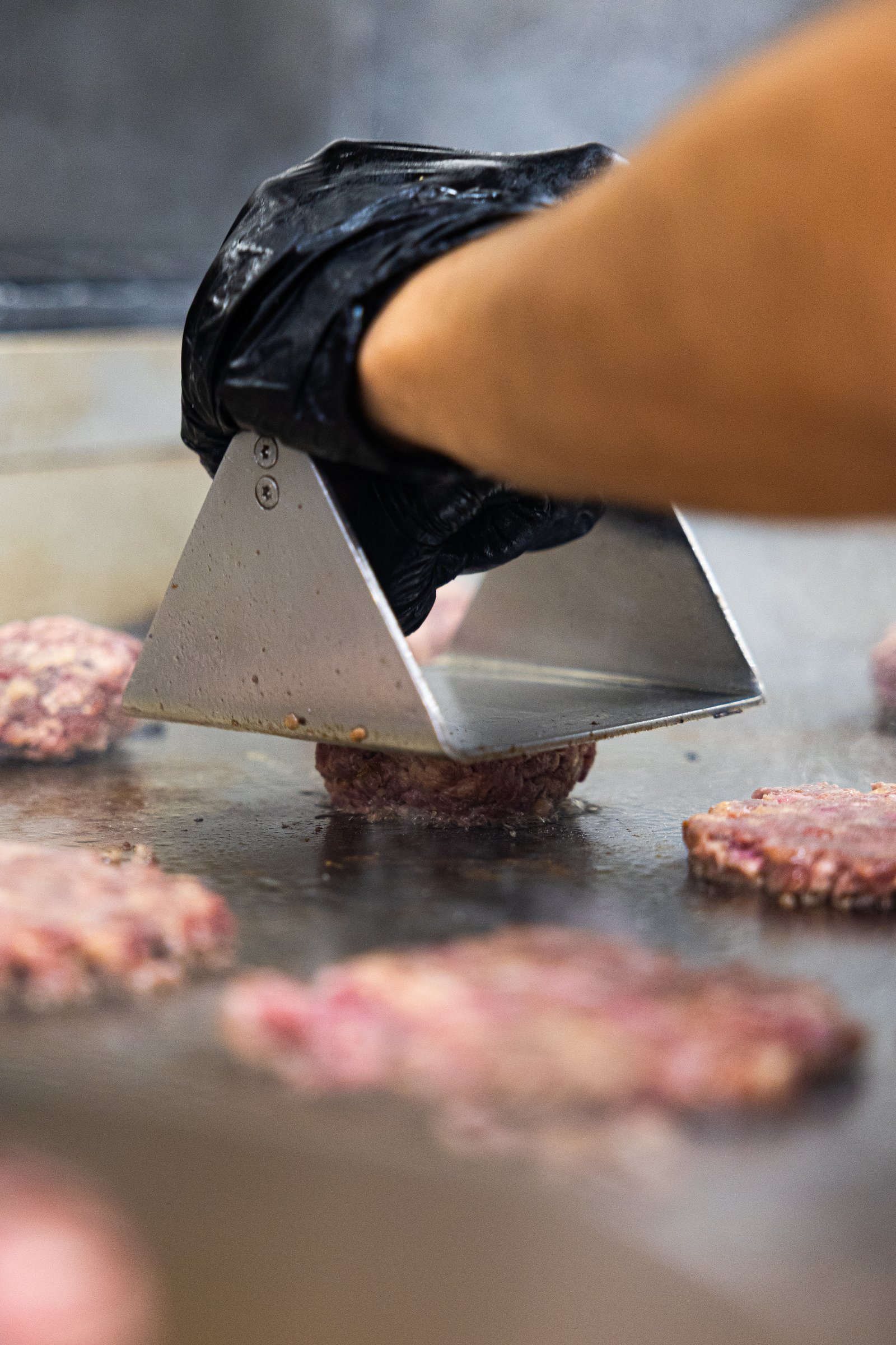 Mano con guante negro presionando una hamburguesa en la plancha