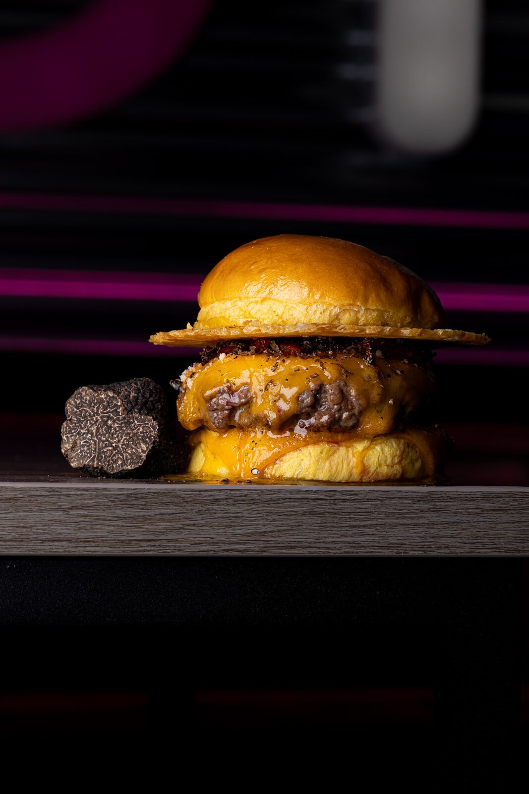 Smash burger con queso fundido y trufa negra sobre tabla de madera con halo magenta — fotografía cinematográfica Vibra Burger