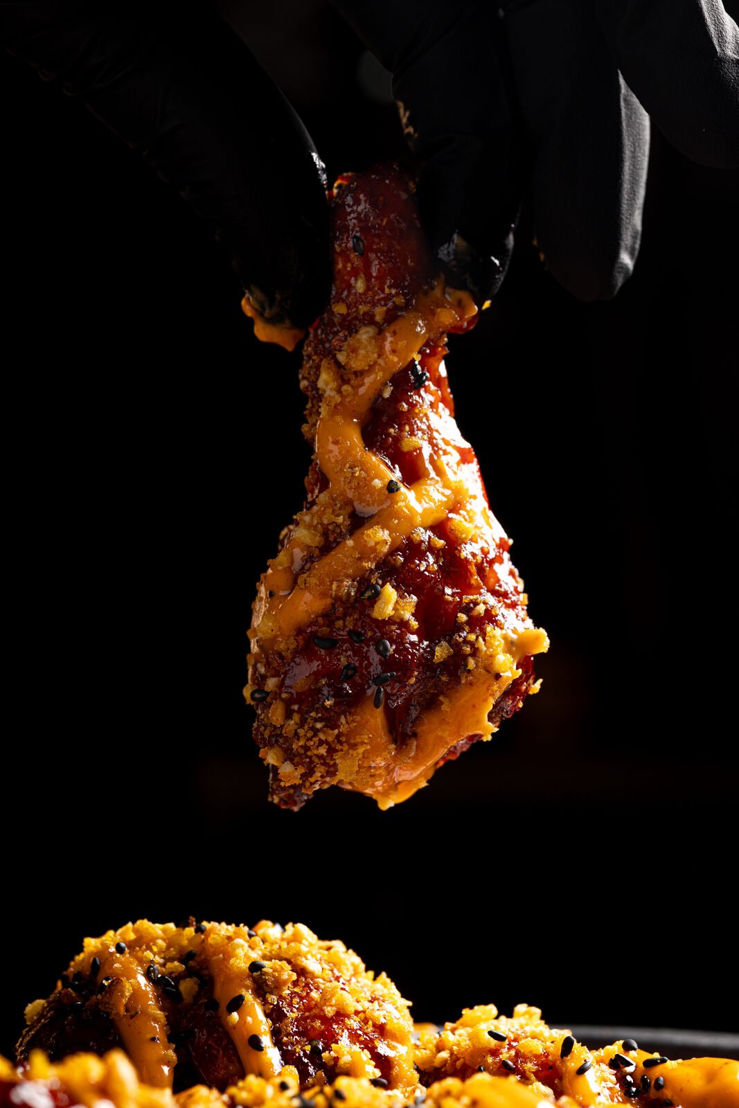 Mano con guante negro sosteniendo un muslo de pollo glaseado con sésamo — primer plano dramático